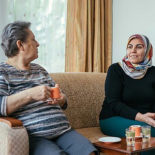 Zwei Frauen sitzen auf einem Sofa und unterhalten sich. Eine trägt Kopftuch.