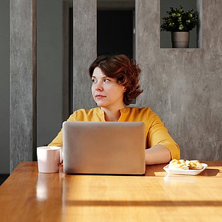 Eine Frau sitzt mit Kaffee und Kuchen vor einem Laptop.